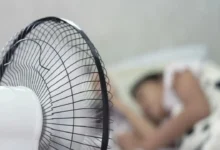 يظهر شخصاً نائماً تحت المروحة مع إبراز المخاطر النوم تحت المروحة الصحية المحتملة مثل جفاف الجلد والعينين ومشاكل التنفس.