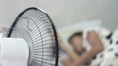 يظهر شخصاً نائماً تحت المروحة مع إبراز المخاطر النوم تحت المروحة الصحية المحتملة مثل جفاف الجلد والعينين ومشاكل التنفس.